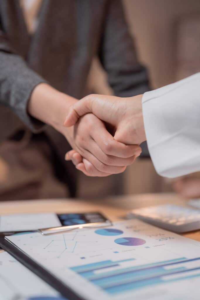 Businesswomen shaking hands after reaching an agreement while working on financial charts and graphs, celebrating successful teamwork and partnership in corporate setting