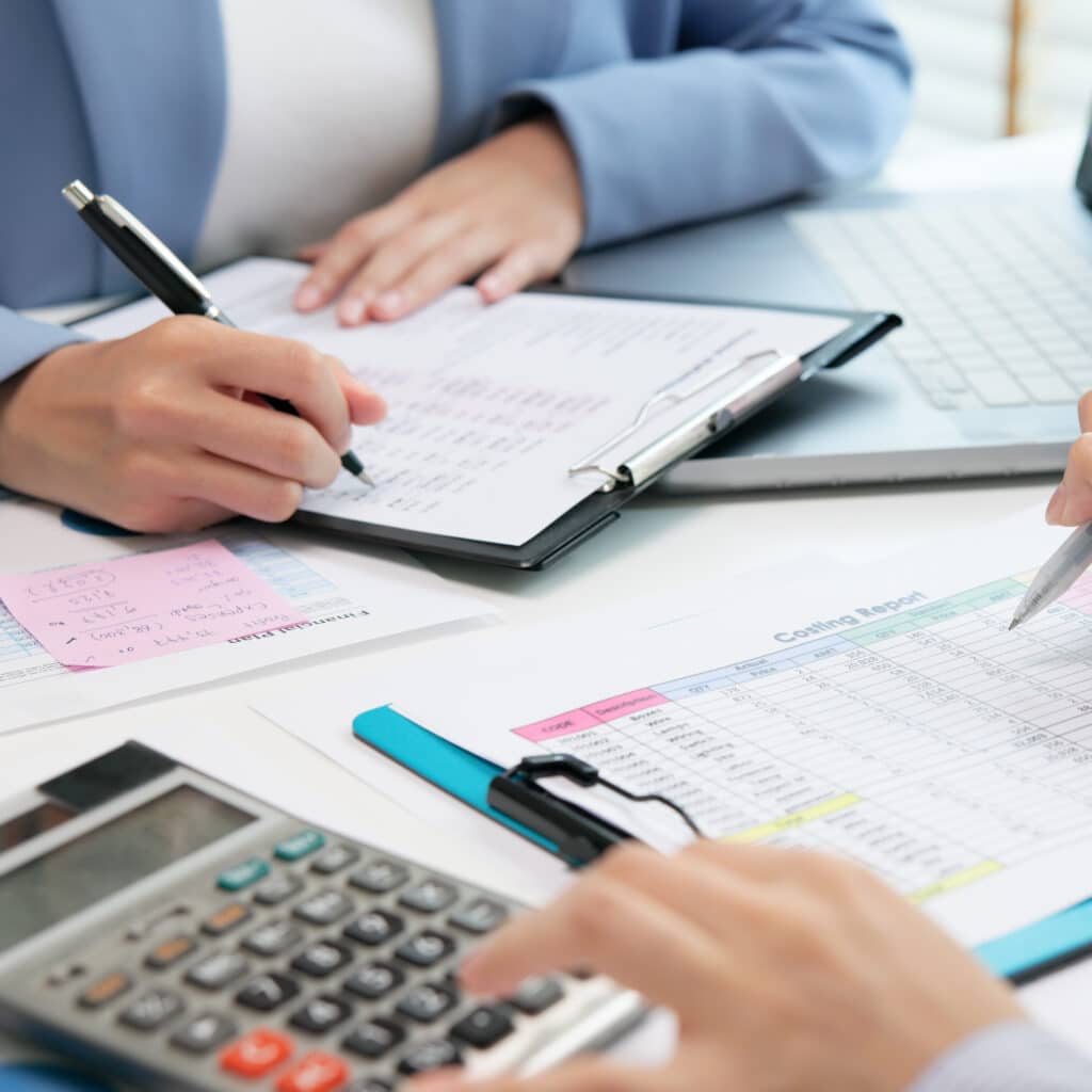Audit et analyse financière : révision des coûts Deux personnes révisent des documents financiers, une écrit sur un presse-papiers et l'autre pointe un 'Costing Report' avec un stylo, près d'une calculatrice.