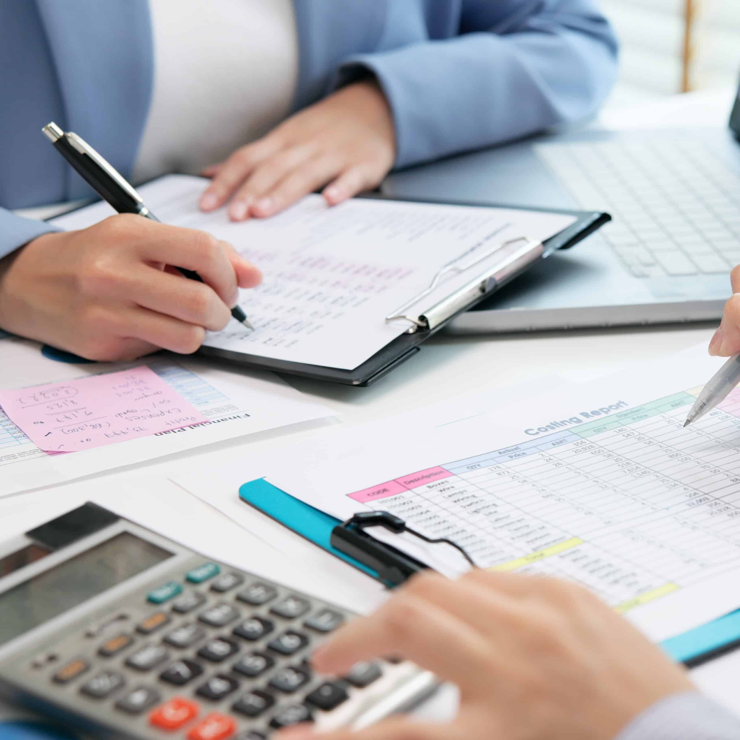 Deux personnes révisent des documents financiers, une écrit sur un presse-papiers et l'autre pointe un 'Costing Report' avec un stylo, près d'une calculatrice.